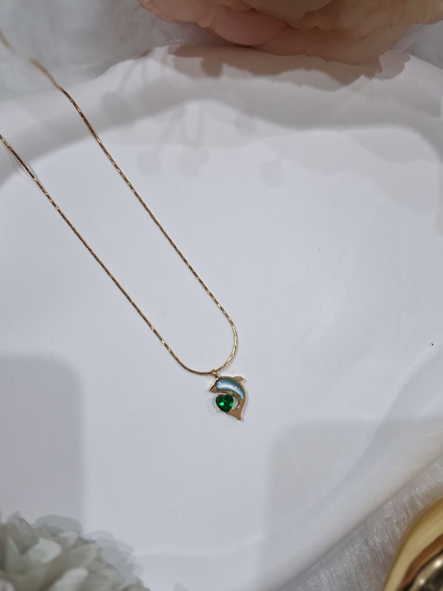 A delicate gold chain necklace with a small crescent moon pendant featuring a green heart-shaped stone, displayed on a white decorative tray with soft pink and white flowers in the background.