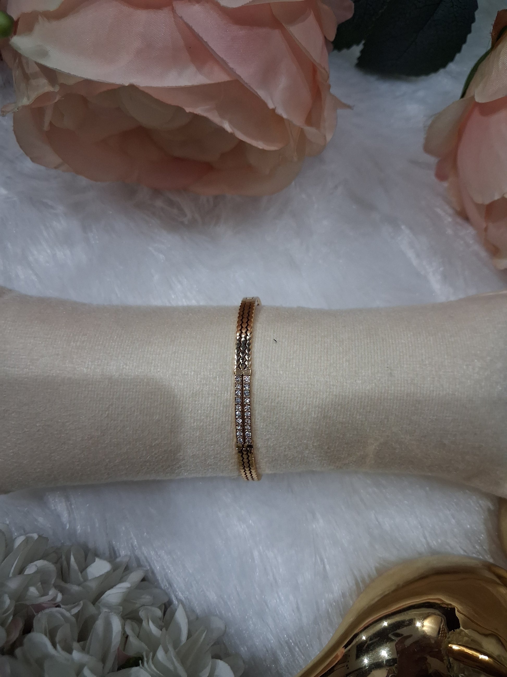 A delicate rose gold bangle bracelet with a row of small clear pavé stones, displayed on a beige fabric jewelry stand. The stand is set against a white faux fur background decorated with pink and white artificial flowers.