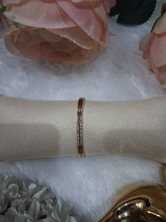 A delicate rose gold bangle bracelet with a row of small clear pavé stones, displayed on a beige fabric jewelry stand. The stand is set against a white faux fur background decorated with pink and white artificial flowers.
